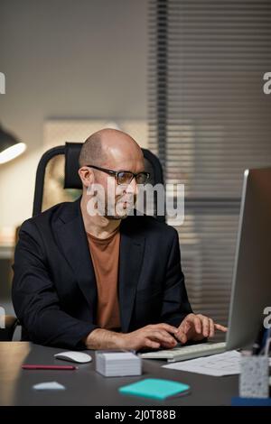 Vertical portrait of bald adult businessman using computer at workplace while working late at night Stock Photo