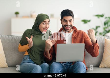 Joyful Muslim Spouses Using Laptop Celebrating Victory Shaking Fists Indoors Stock Photo