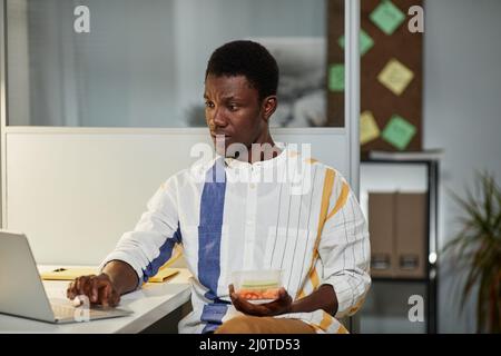 Portrait of young African American man using laptop while working in office late with healthy snack, copy space Stock Photo