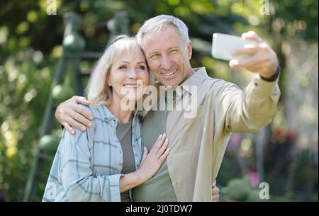 Happy elderly couple spending time in their garden together, taking selfies and smiling at cellphone camera Stock Photo