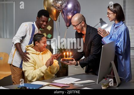 Diverse group of colleagues bringing Birthday cake with candles surprising young woman in office Stock Photo