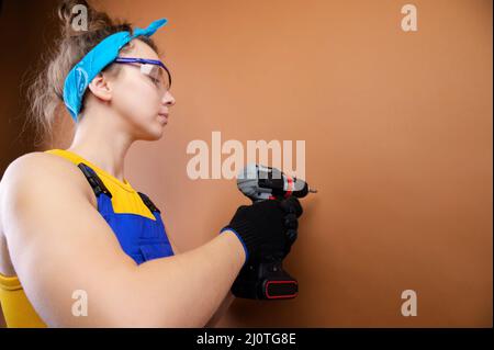 Young woman repairman in overalls and goggles works with a screwdriver in the studio on a brown background. Focus on the screwdr Stock Photo