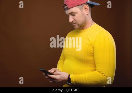 Pleasant caucasian mature casual man uses phone smartphone in studio. Using electronics, typing SMS and surfing the Internet Stock Photo