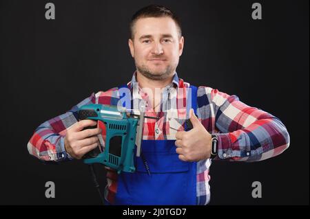 Portrait of a friendly carpenter with an electric jigsaw in his hand showing a thumb up. Studio shot on black background Stock Photo