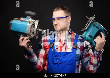 A professional carpenter in uniform and goggles holds an electric tool, a jigsaw and a milling cutter. Studio portrait on black