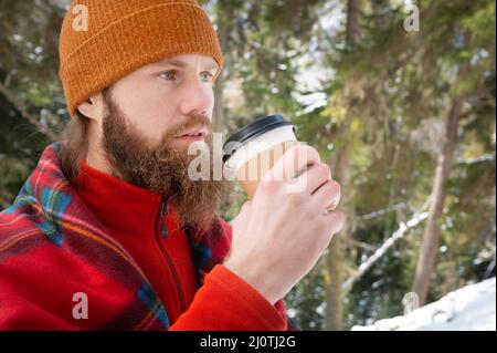 Bearded caucasian man in a hat wrapped in a blanket with a mug of hot drink in a paper cup in a winter coniferous forest on a su Stock Photo