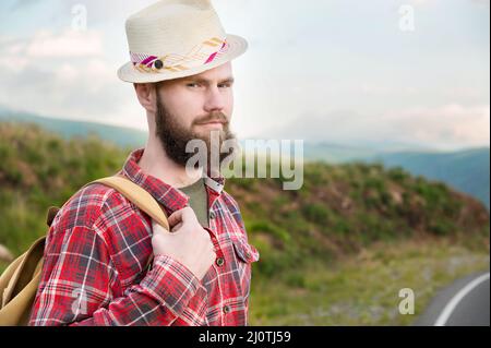 Attractive romantic bearded Caucasian man wearing a hat, plaid shirt and a backpack. Stands in the countryside in the mountains Stock Photo