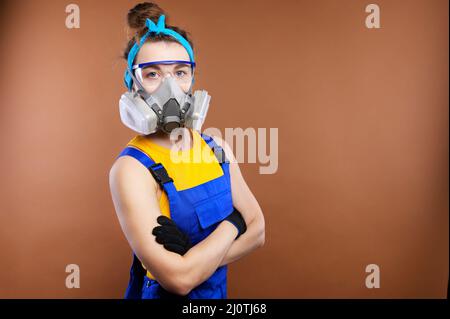 Portrait of Caucasian young woman builder. The arms are folded over the chest. Wearing protective goggles and a respirator weari Stock Photo