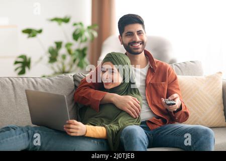 Middle Eastern Spouses Using Laptop And Watching Television At Home Stock Photo