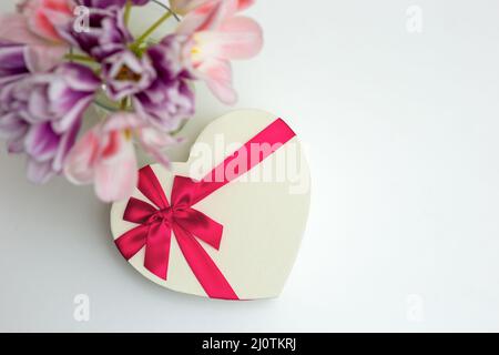 A bouquet of open pink and purple tulips , a heart-shaped gift box, with a pink bow, on a white table . Stock Photo