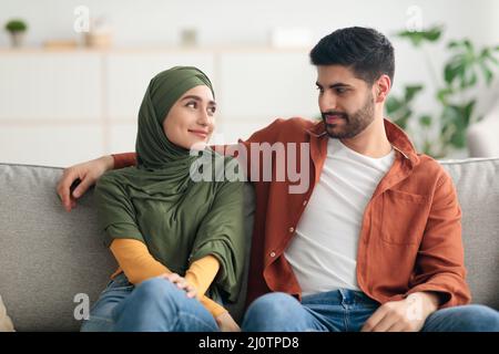 Happy Muslim Wife Wearing Hijab Looking At Husband Sitting Indoors Stock Photo