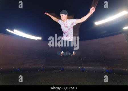 A young skateboarder rides at speed sideways along the ramp by spreading his hands to the sides. Night shot with long exposure a Stock Photo
