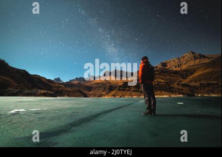 A brooding man stands on the surface of a frozen lake illuminated by moonlight high in the mountains against the backdrop of epi Stock Photo