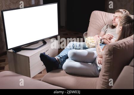 Attractive young long-haired couple hugging on a sofa at home in isolation watching tv. The concept of spending time with loved Stock Photo