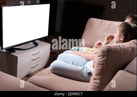 Attractive young long-haired couple hugging on a sofa at home in isolation watching tv. The concept of spending time with loved Stock Photo