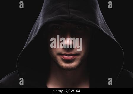 Close-up portrait of a courageous man in a deep dark hood on a black background. The concept of secrecy of secrets and people hi Stock Photo