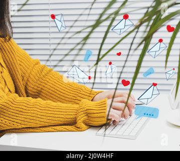 A woman sits at a white table and types on a wireless keyboard, letters and hearts are flying. Social media addiction, spam and Stock Photo