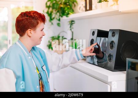 Modern, short-haired, red-haired, non-binary woman, turns on mini-system at home. Stock Photo