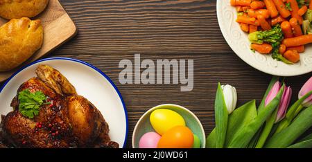 Easter food on dark rustic table: pastel colored eggs, roasted chicken, vegetables, buns Stock Photo