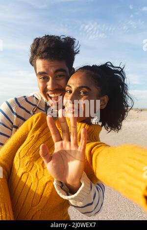 Portrait of smiling biracial woman taking selfie with boyfriend gesturing stop sign at beach Stock Photo