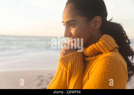 Thoughtful young biracial woman wearing yellow sweater looking away at beach during sunset Stock Photo