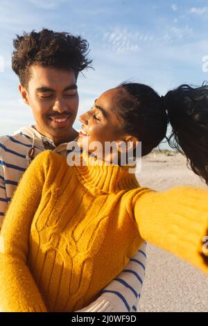 Young biracial woman taking selfie with smiling boyfriend at beach against sky on sunny day Stock Photo