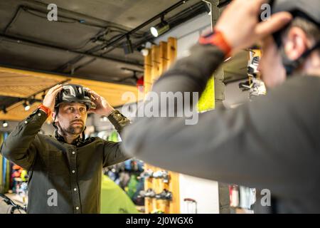 Young man came to the bicycle store. He is measuring the helmet. Male chooses helmet in sports equipment store. Purchase of new Stock Photo
