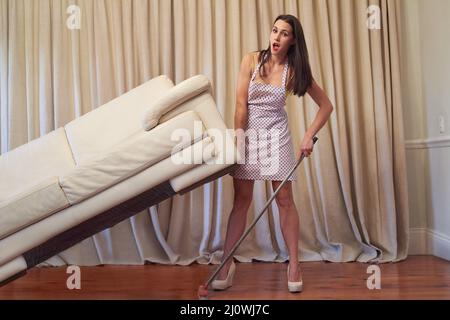Keeping the house clean takes super strength. Portrait of a young woman lifting a couch and sweeping the floor beneath it. Stock Photo