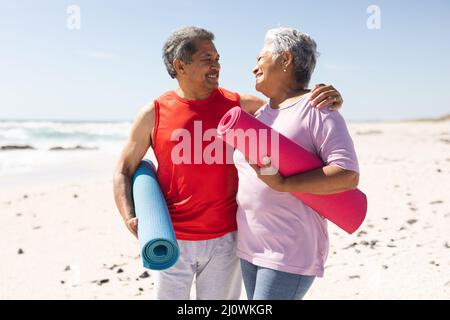 Smiling senior biracial couple looking at each other holding rolled yoga mats on sunny beach Stock Photo