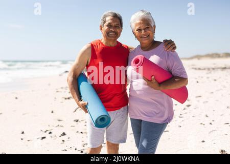 Portrait of smiling senior biracial couple holding yoga mats at beach on sunny day Stock Photo