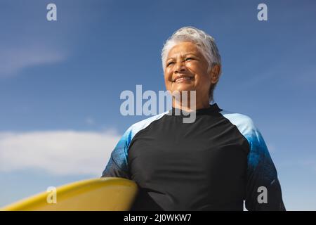 Low angle view of smiling senior biracial woman carrying surfboard against blue sky on sunny day Stock Photo