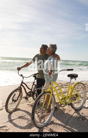 Multiracial senior couple looking away standing with bicycles at beach against sky during sunny day Stock Photo