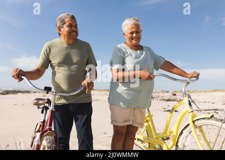 Happy multiracial senior couple standing with bicycles at beach against sky during sunny day Stock Photo