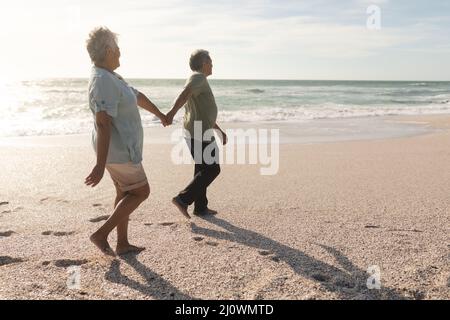 Full length side view of multiracial senior couple holding hands while walking on shore at beach Stock Photo