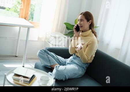 Young Asian woman talking with friend by cell phone while sitting on sofa at home. Stock Photo