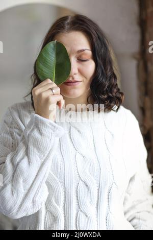 Girl with beautifully styled hair in white sweater, calmly stands covering one eye with green leaf Stock Photo