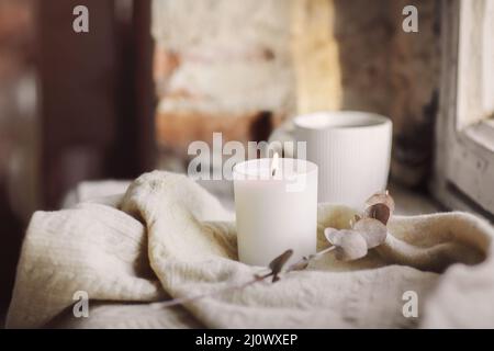 White burning candle on woolen plaid and cup of tea on windowsill at home in cozy atmosphere Stock Photo