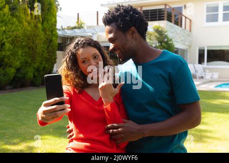 Smiling young multiracial couple showing engagement ring while taking selfie on smartphone in garden Stock Photo