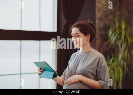 Young smiling pregnant woman holding digital table and making video call while sitting on bed at home, talking online with famil Stock Photo