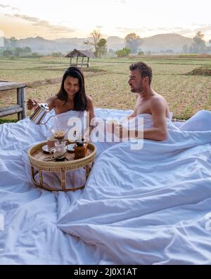 Sunrise over rice paddies in Nan Thailand, man and woman in outdoor bed waking up with coffee in the morning in Thailand Nan Stock Photo