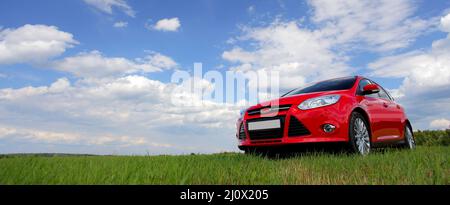 Red car on a blue sky background with white clouds. The car is standing in the field on the green grass. Stock Photo