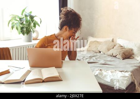 Tired flustrated African descent young girl sitting at desk in front of laptop while yawning cover mouth with hand not having in Stock Photo