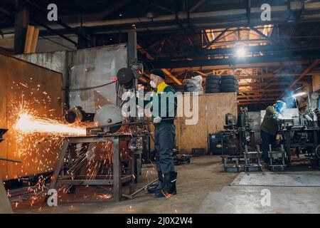 Workers working with metal construction on plant. Metal processing with big angle grinder disk saw and welder welding. Sparks in Stock Photo