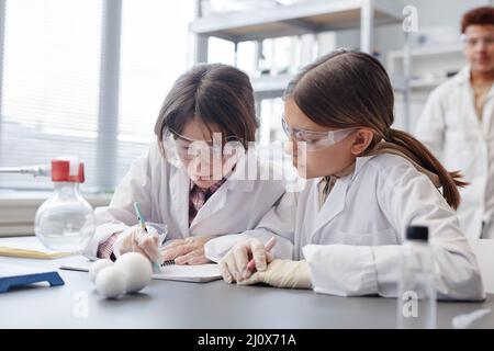 Portrait of two girls taking notes in notebook while doing science experiment in chemistry lab at school Stock Photo