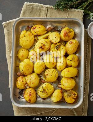 Oven baked whole crushed and crusty potato spuds with seasoning and herbs in metalic tray Stock Photo