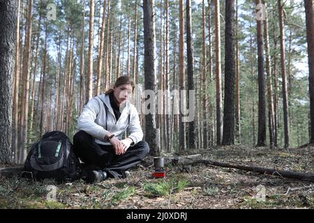 A man in a campaign drinks tea. A young traveler in the woods camped and cooked breakfast in nature. The naturalist drinks hot fresh tea cooked on ope Stock Photo