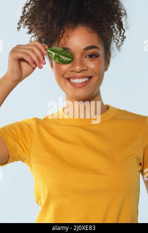 Natural beauty. Happy Afro american girl in yellow tshirt with curly hair covering one eye with green leaf. Stock Photo