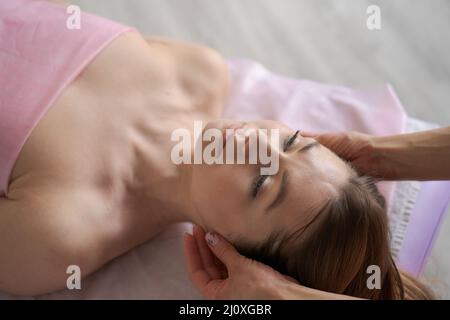 Woman lay on couch on her back with her eyes closed and enjoy. Makes relaxing and therapeutic head massage with weight. Spa client threw his head back and rejuvenate. Wellness treatments in SPA Stock Photo