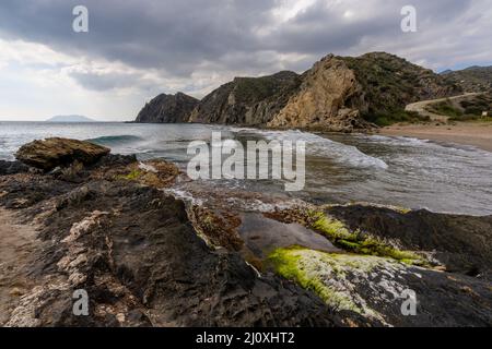 A secluded sandy beach on a wild mountainous coastline with colorful rocks and algae in the foreground Stock Photo