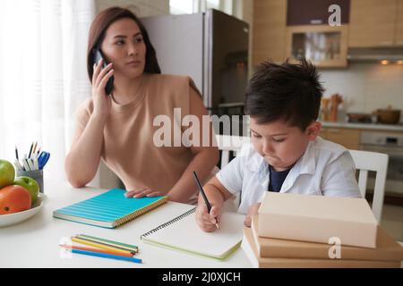 Schoolboy doing homework under control of his mother talking on phone with teacher Stock Photo